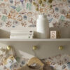 CHARLOTTE LIGHT wallpaperwith a selection of shells, seaweed, starfish and seahorses in blue, beige, green being nused in a childrens room with a cream shelf unit with biooks and toys on it and a beige jumper on a hanger