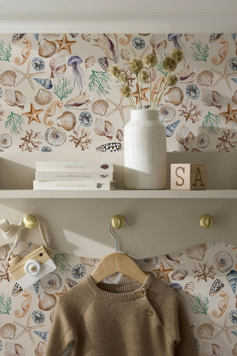 CHARLOTTE LIGHT wallpaperwith a selection of shells, seaweed, starfish and seahorses in blue, beige, green being nused in a childrens room with a cream shelf unit with biooks and toys on it and a beige jumper on a hanger