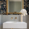 CHARLOTTE MIDNIGHT wallpaper dark background with a selection of shells, seaweed, starfish and seahorses in blue, beige, green used in a bathroom above a tiled section above a basin with a gold ornate mirror and vase with fake flowers
