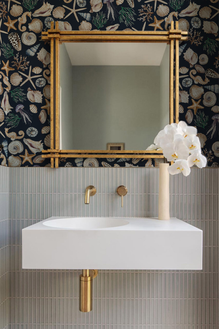 CHARLOTTE MIDNIGHT wallpaper dark background with a selection of shells, seaweed, starfish and seahorses in blue, beige, green used in a bathroom above a tiled section above a basin with a gold ornate mirror and vase with fake flowers