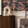 MINA MIDNIGHT wallpaper with dark background and orange, pink and green fern designs on the wall above a panelled area with a dark wood side board and chair and coffee table in front. Mina is part of our home decor collection The Series Collection. Contact Curate Space for more details